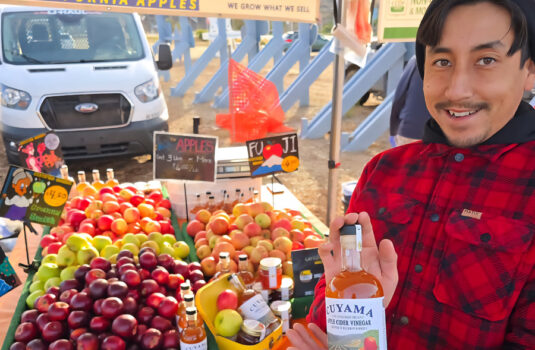 Man standing in front of a table full of all different types of apples. He holds up apple cider vinegar.