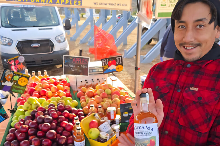Man standing in front of a table full of all different types of apples. He holds up apple cider vinegar.