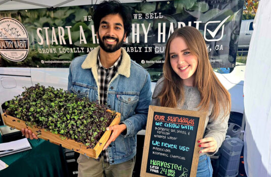 Two people, a man and a women stand holding microgreens and a sign listing their practices.