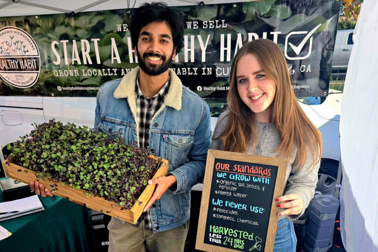 Two people, a man and a women stand holding microgreens and a sign listing their practices.