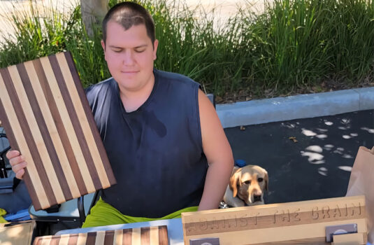 Louie the owner and master carver of Against the Grain stands with his creations: an array of wooden cutting boards. He hold a striped cutting board in his hands. Hi Louie!