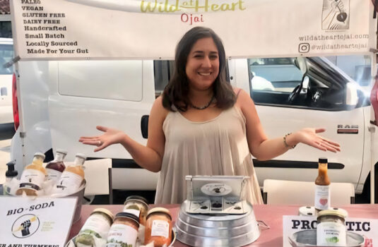 Michelle from Wild at Heart, stands smiling with her hands out. She is behind a table full of her probiotic products.