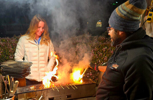 A man and a woman stand on either side of a charcoal fire.