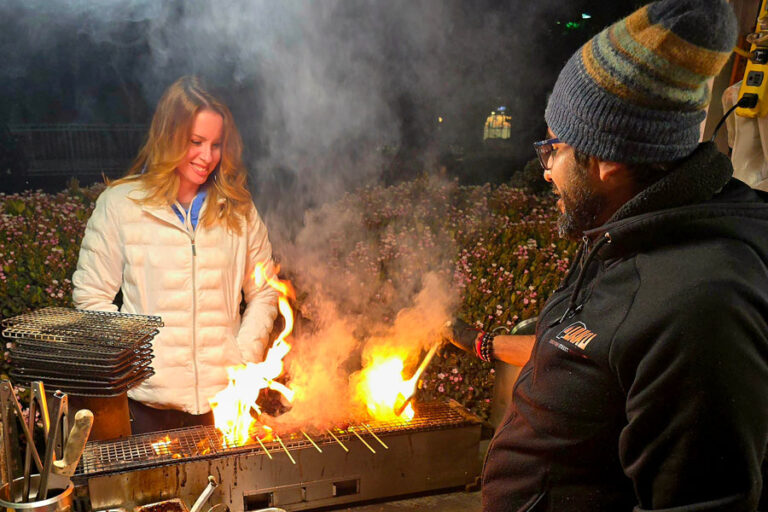 A man and a woman stand on either side of a charcoal fire.